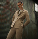 A person wearing a beige suede blazer and matching trousers, standing in front of a corrugated metal wall.