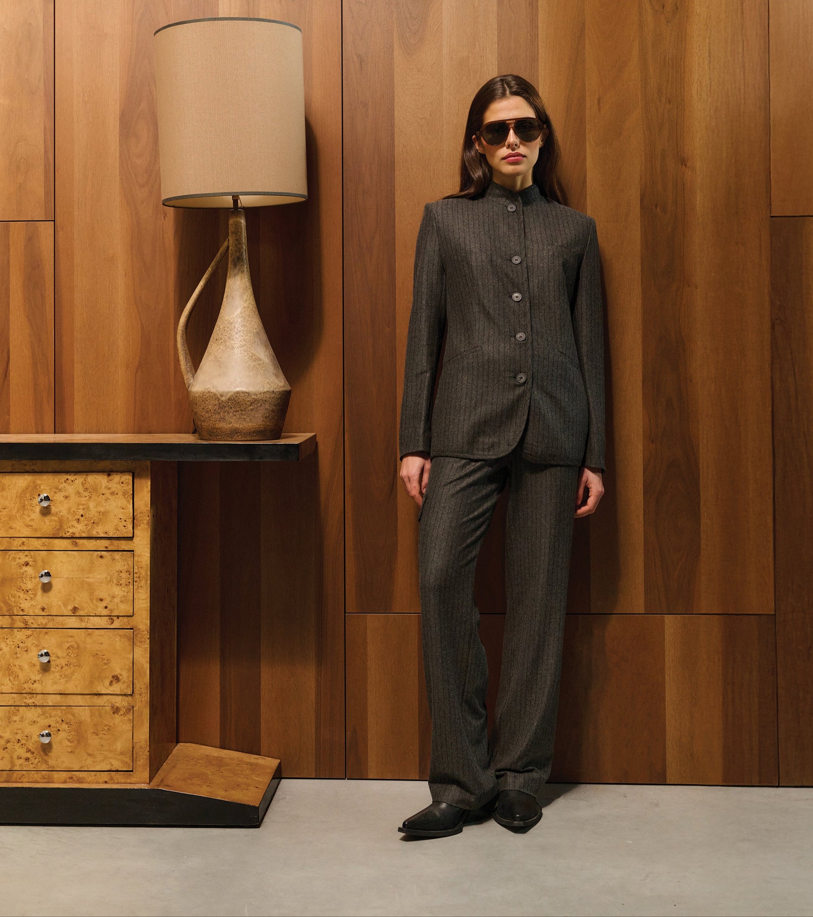 Woman in a gray suit standing against a wooden wall with a lamp and dresser.